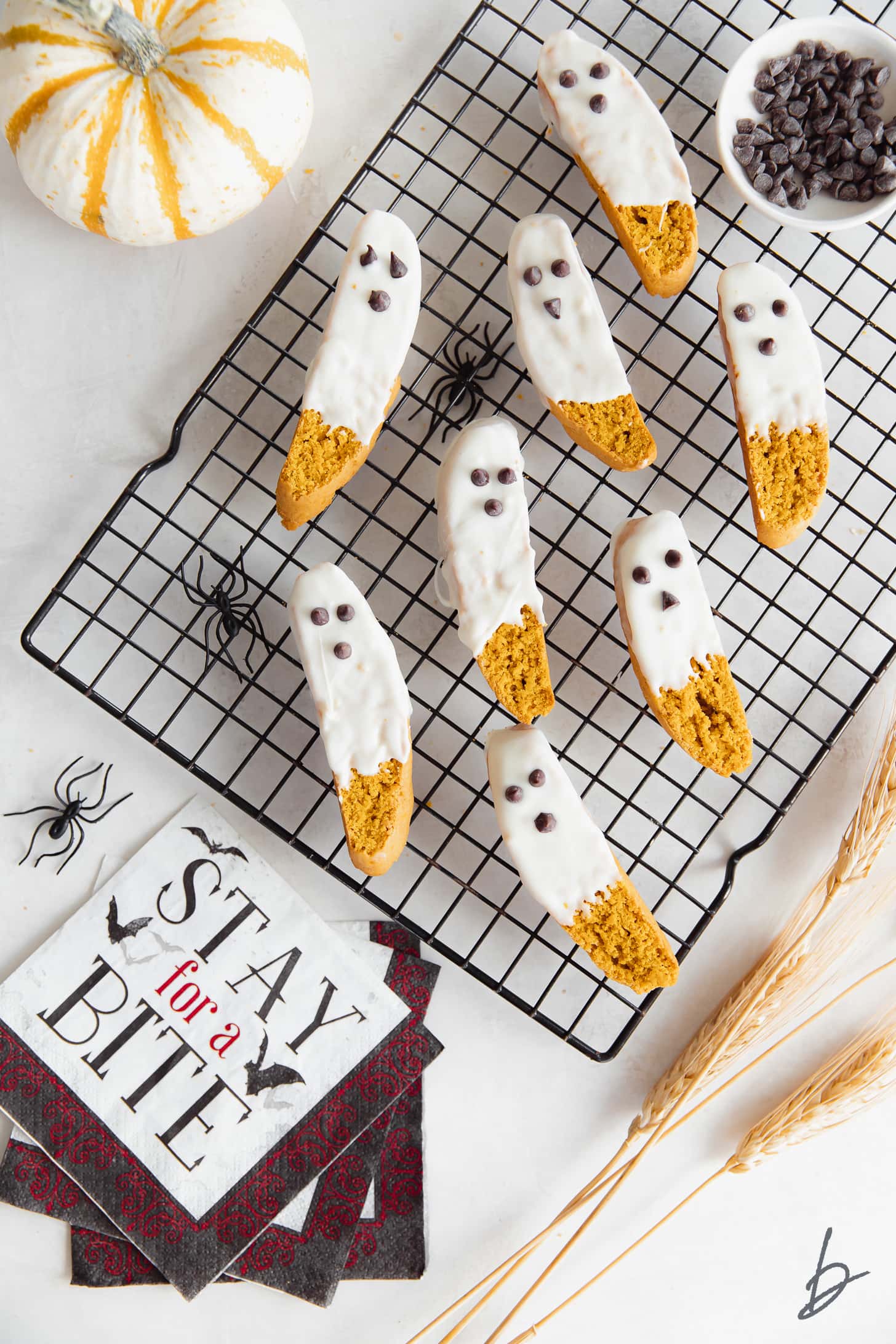 White chocolate dipped pumpkin biscotti that look like ghosts on black wire cooling rack next to halloween napkin and mini pumpkin.