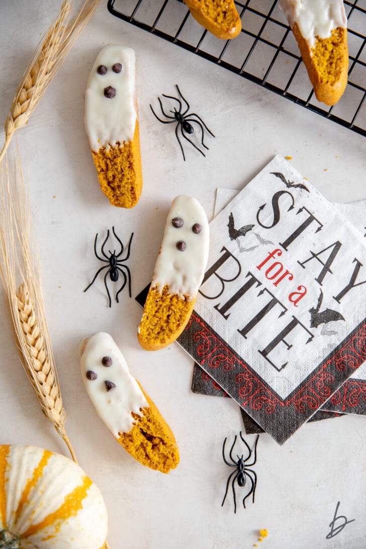 three white chocolate dipped pumpkin biscotti that look like ghosts next to fake spiders and halloween napkin