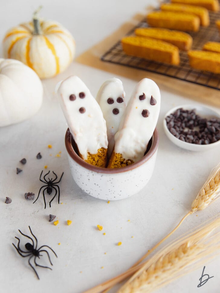 a cup with three white chocolate dipped pumpkin biscotti decorated to look like ghosts