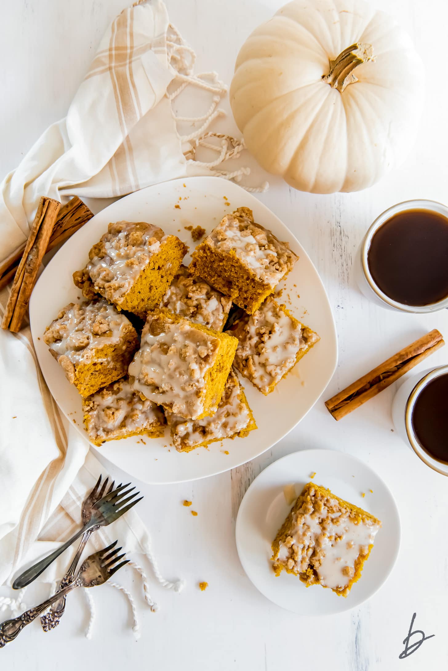 pumpkin coffee cake squares on white round plate next to white pumpkin and mugs of coffee