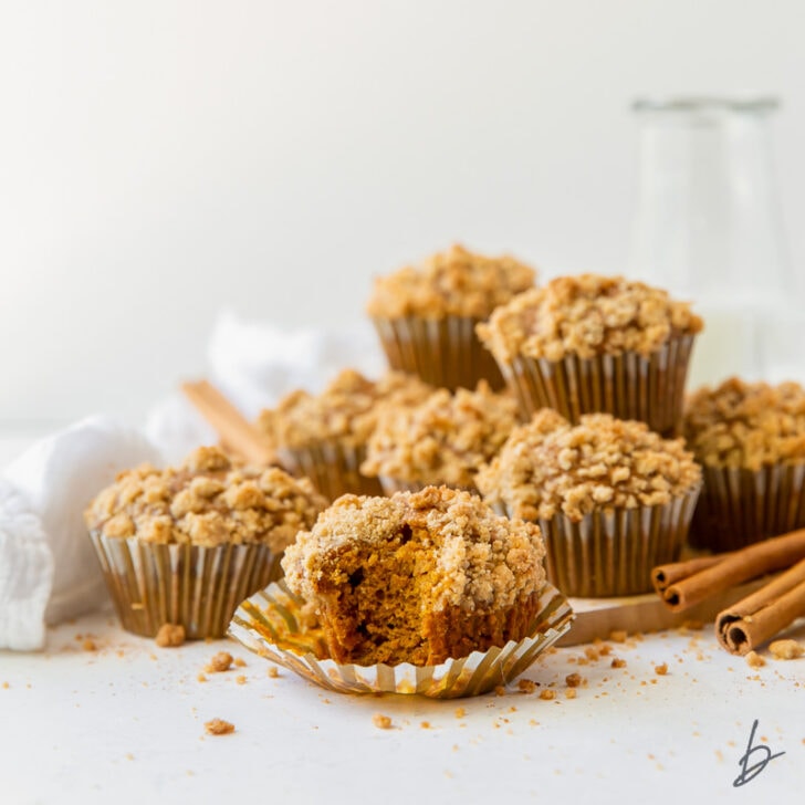 pumpkin muffins with streusel in a pile with front muffin with a bite sitting on open muffin paper liner