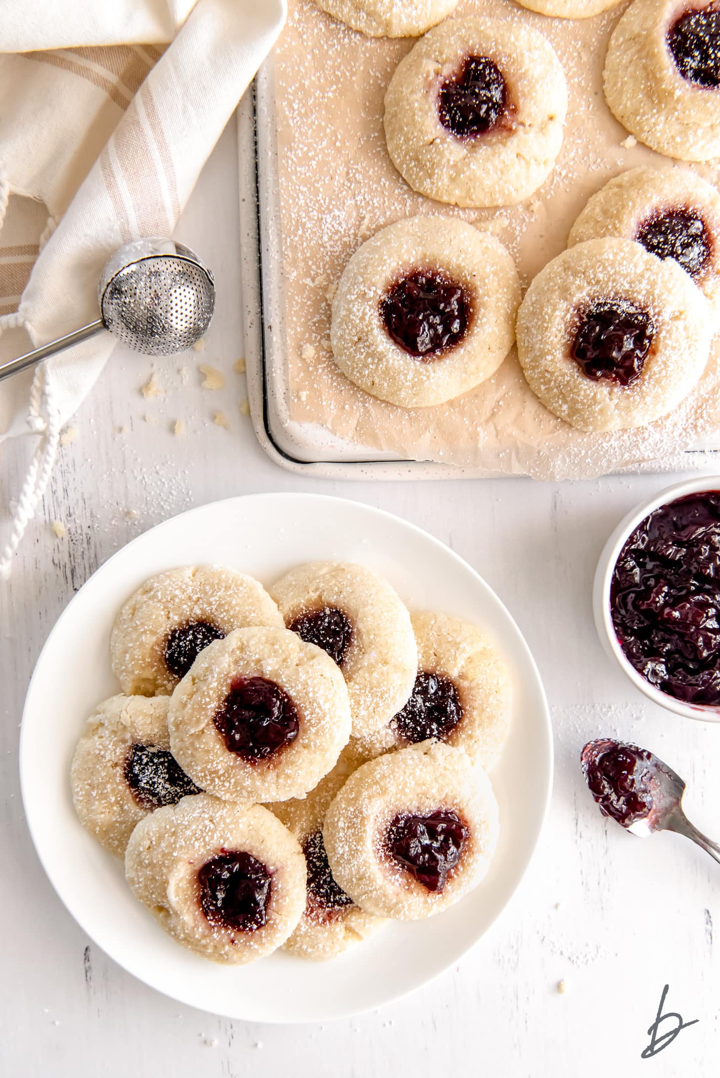 jam thumbprint cookies on a round plate next to bowl of jam and parchment paper with more cookies