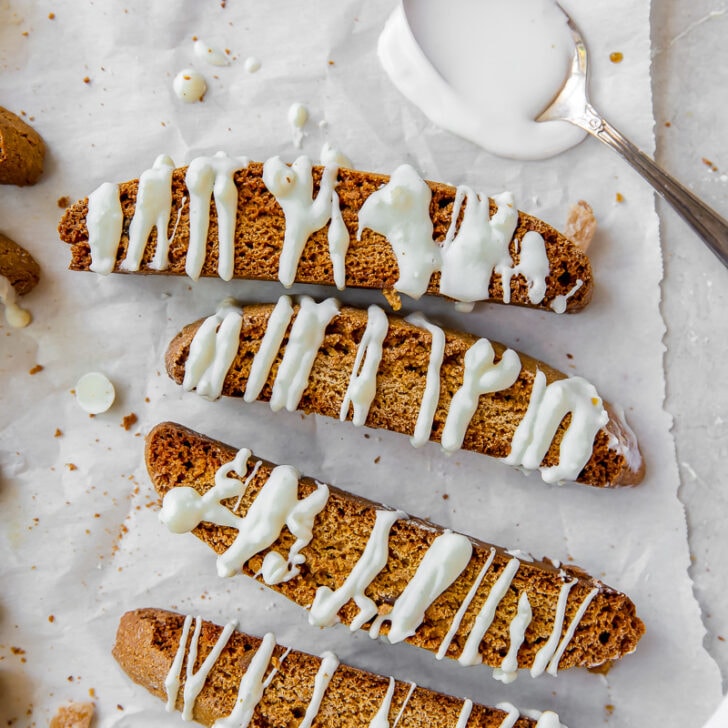 white chocolate drizzled gingerbread biscotti on white parchment paper next to spoon with white chocolate