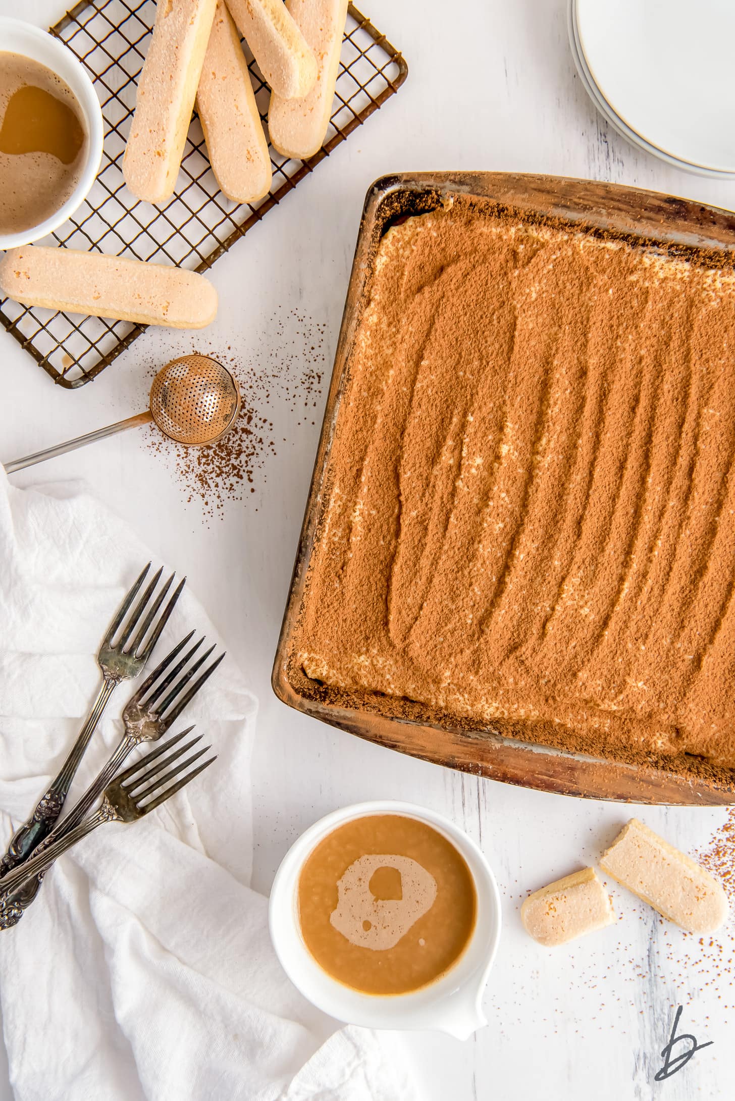 cocoa dusted tiramisu in a square pan next to mug of coffee, a few forks and ladyfingers