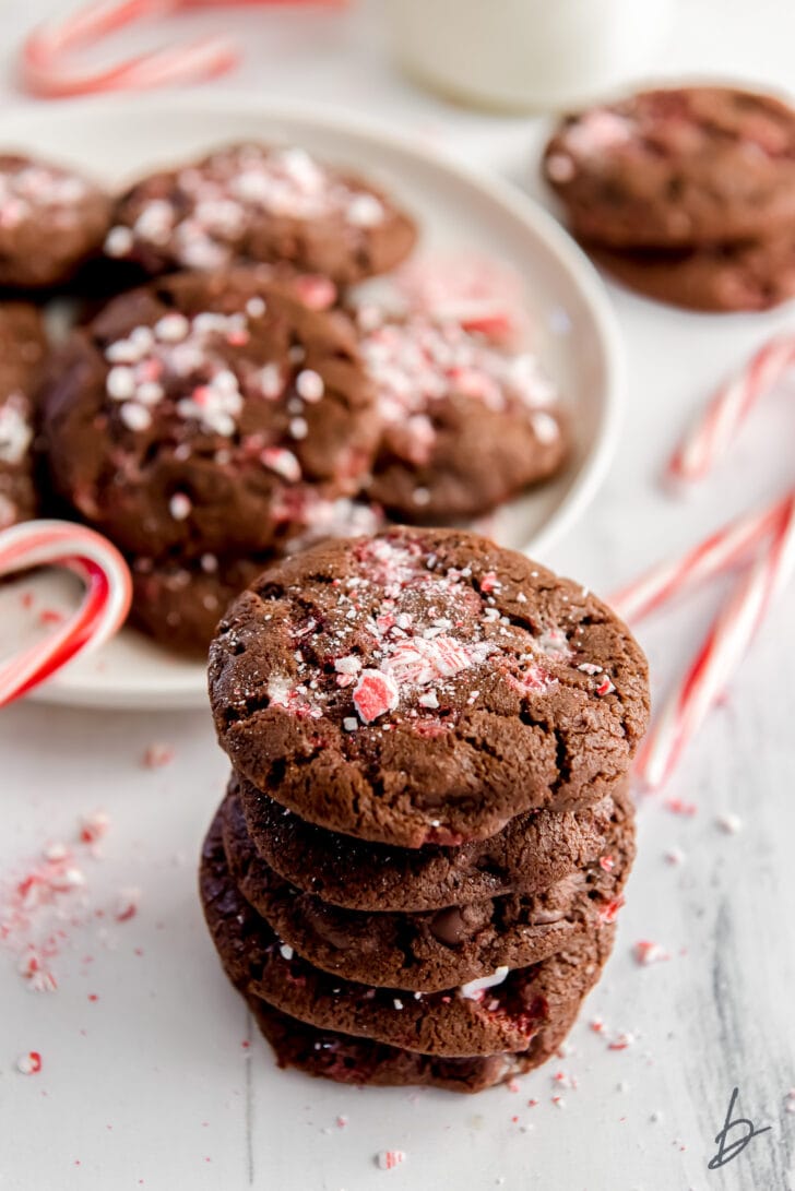 stack of four double chocolate peppermint cookies with crushed peppermint on top in front of plate with more cookies