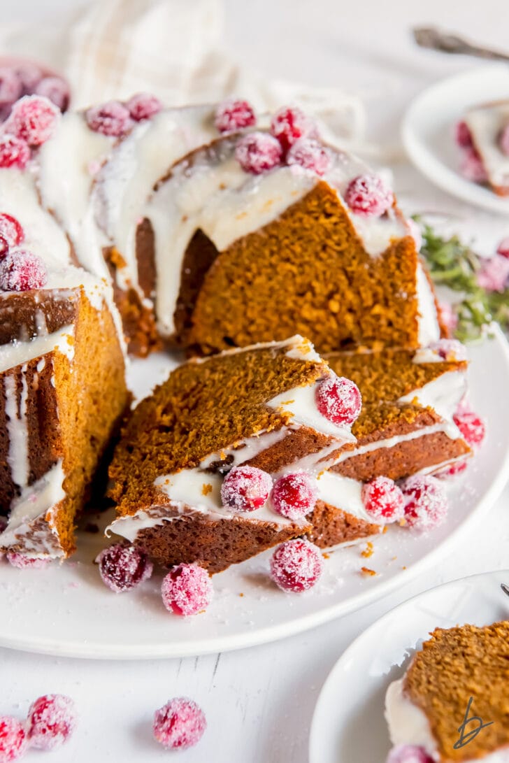 slices of gingerbread bundt cake leaning up against each other and full cake with cream cheese frosting and sugared cranberries