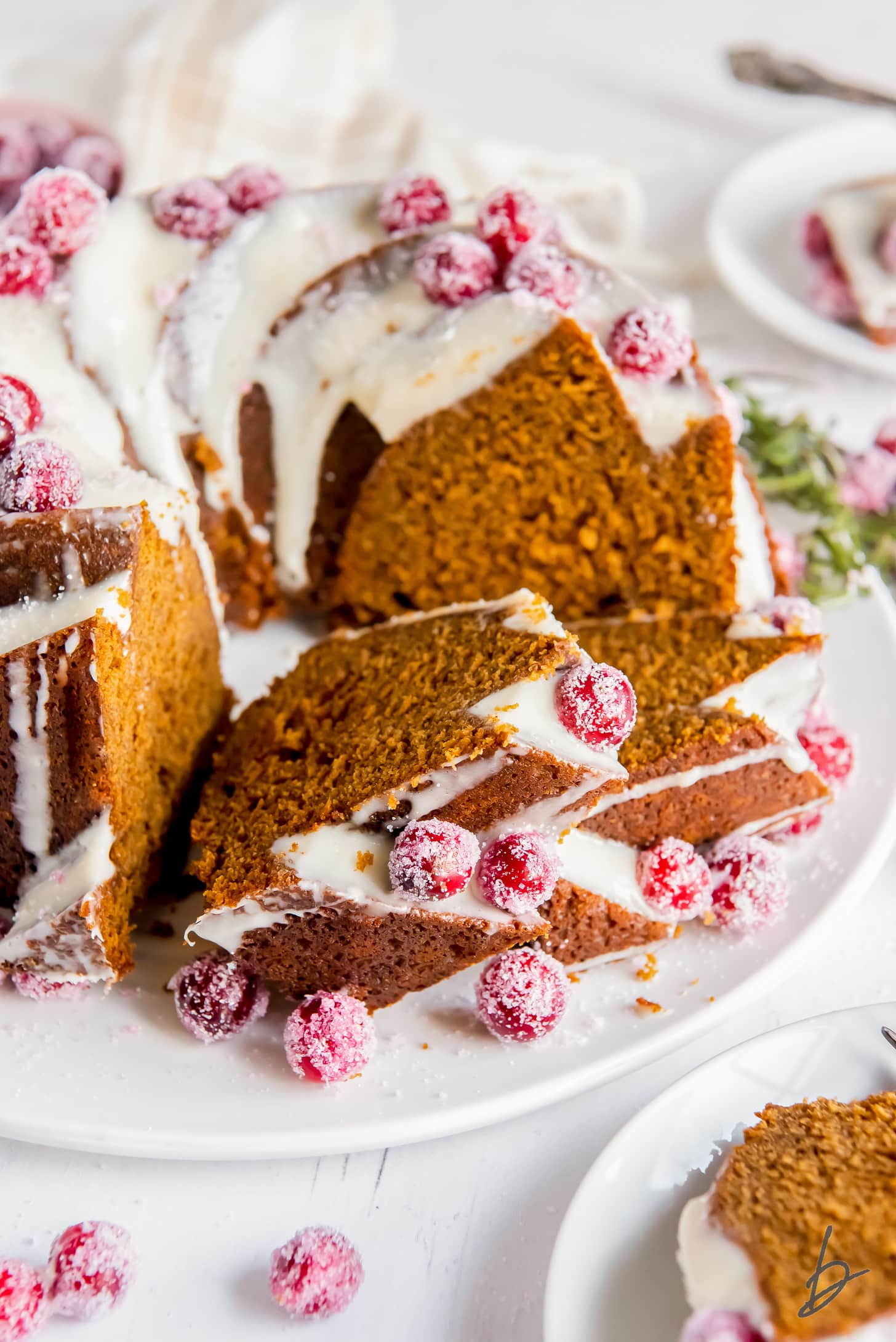 slices of gingerbread bundt cake leaning up against each other and full cake with cream cheese frosting and sugared cranberries.