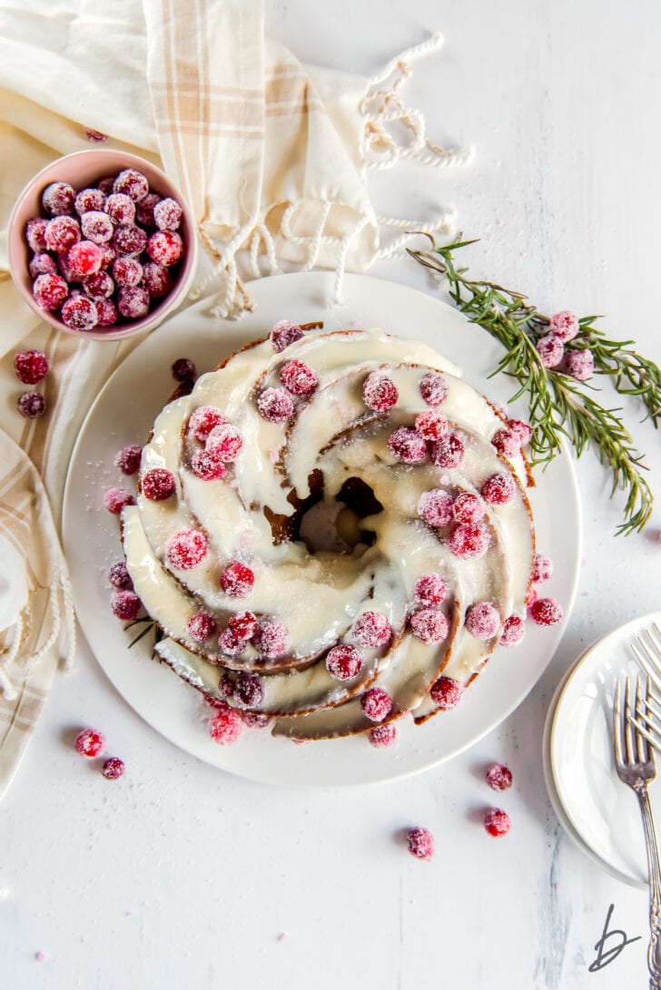 gingerbread bundt cake topped with cream cheese frosting and sugared cranberries next to bowl of cranberries and rosemary sprig