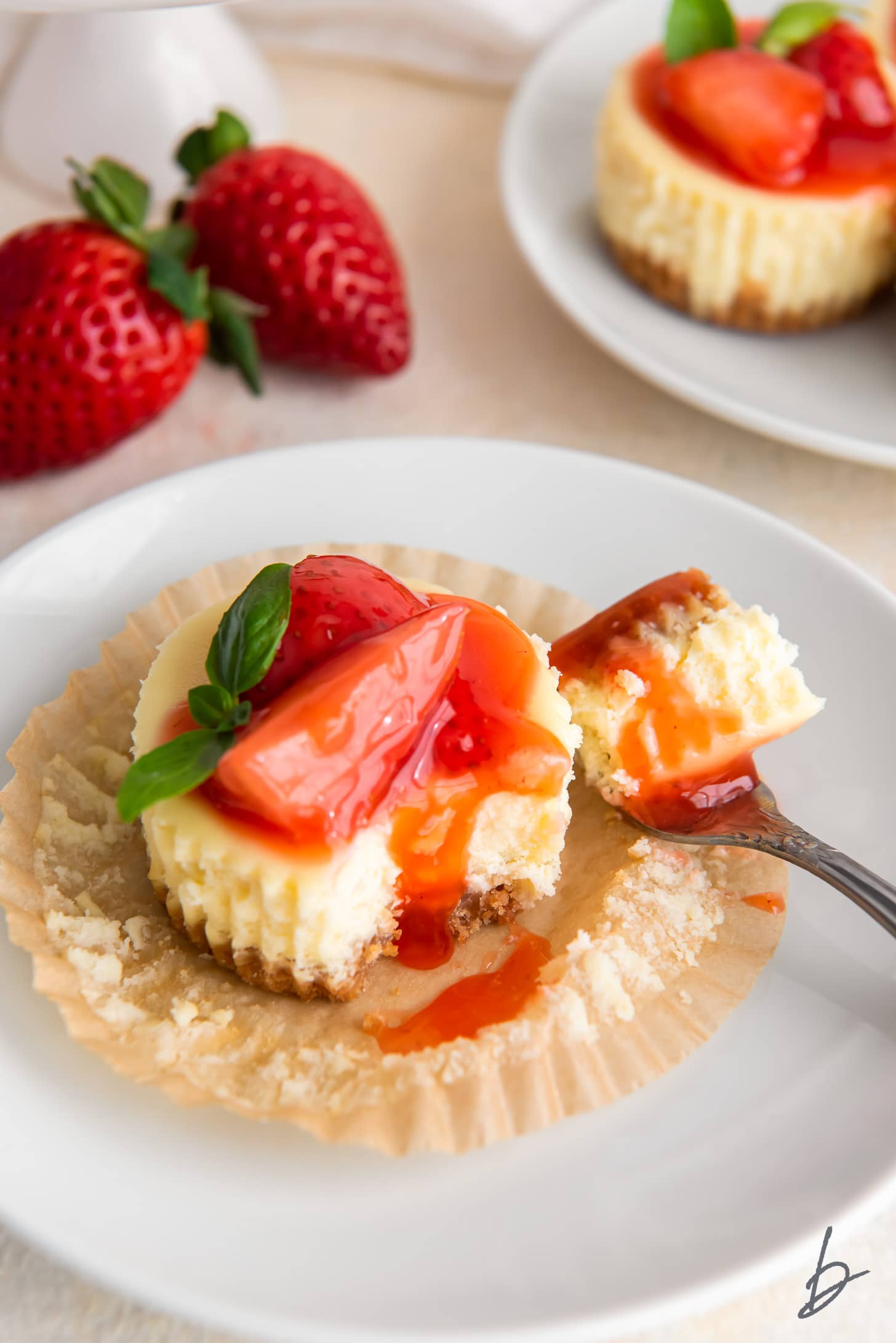 fork taking bite of mini cheesecake with strawberry sauce on paper muffin liner.