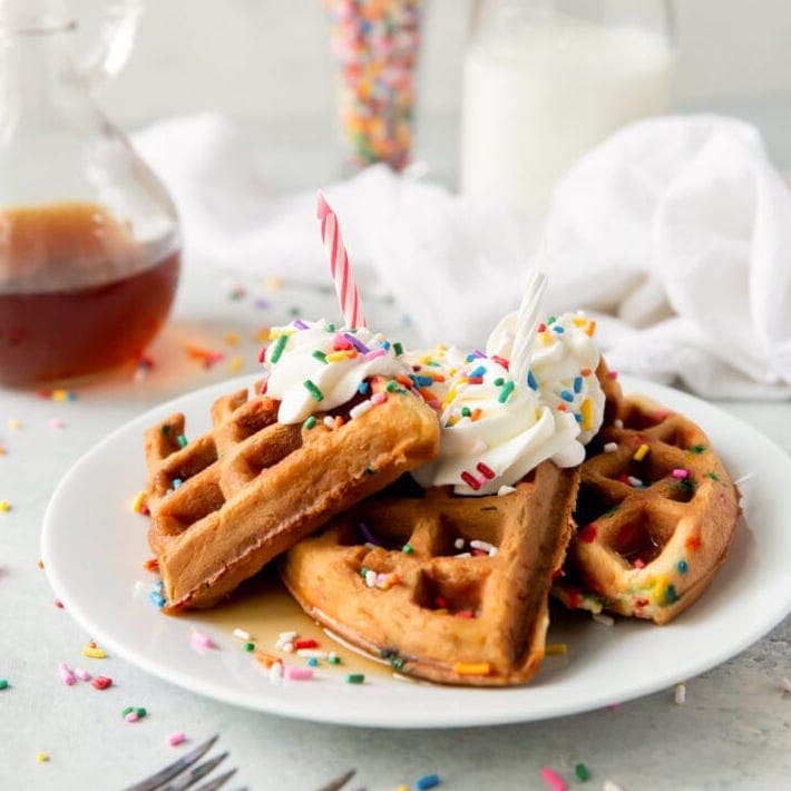 stack of waffles with sprinkles, whipped cream and a pink birthday candle