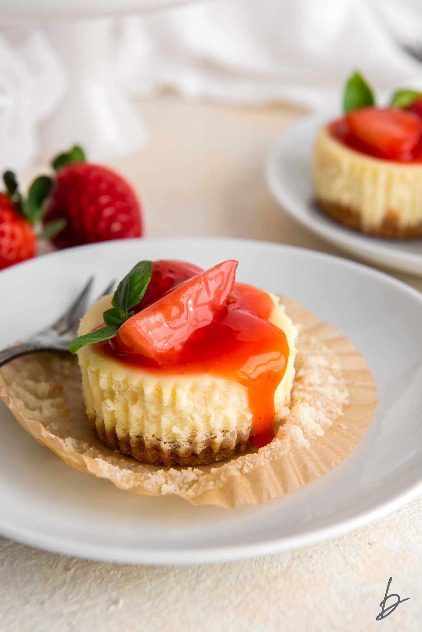 mini cheesecake with strawberry sauce on open paper muffin liner on white plate.