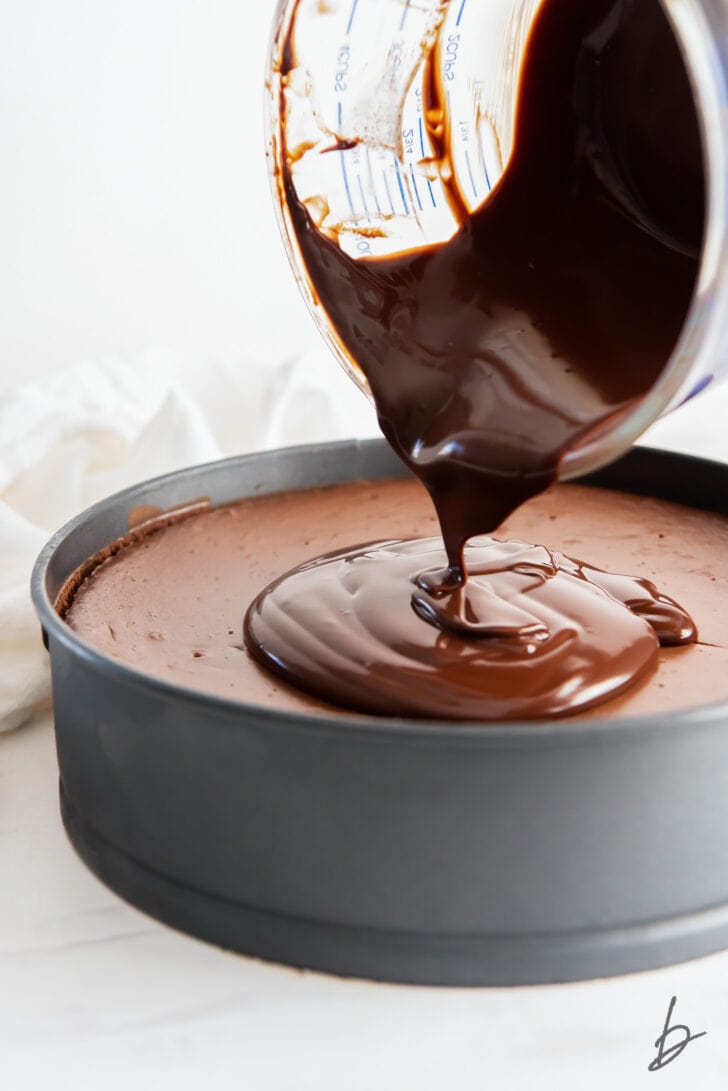 chocolate ganache being poured on top of chocolate cheesecake in a springform pan