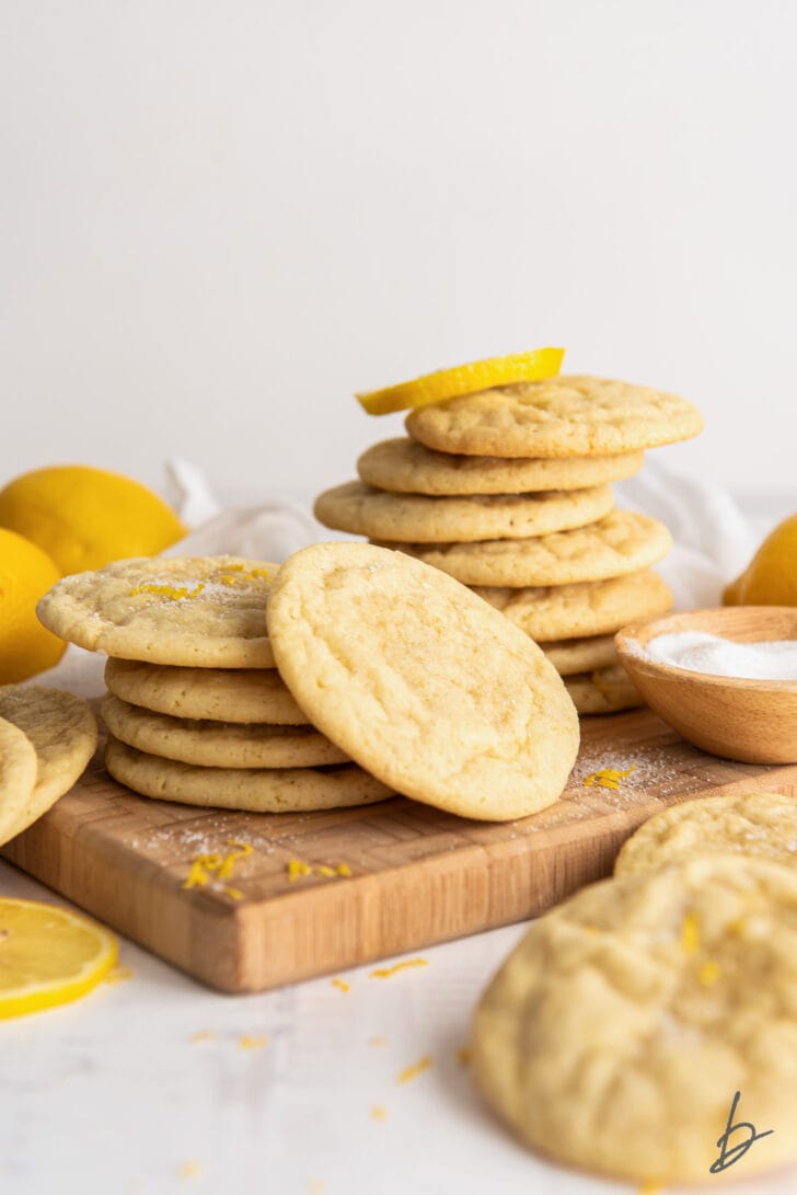 lemon sugar cookies piled on a wood cutting board in front of lemon halves