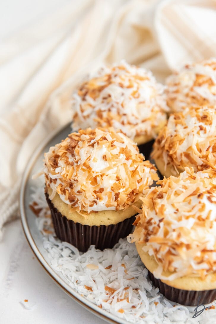 coconut cupcakes garnished with toasted coconut on a plate next to a kitchen cloth