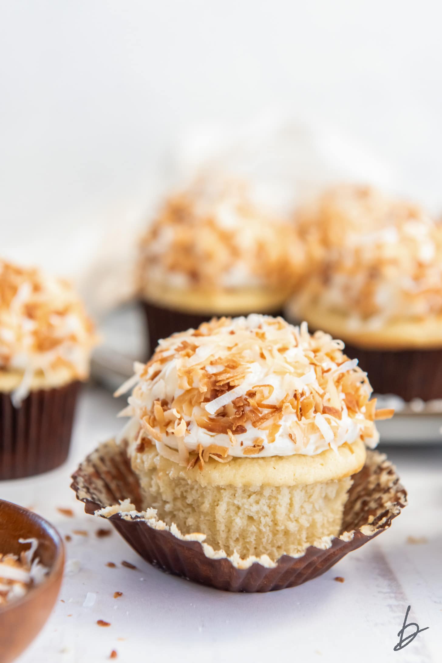 coconut cupcake with frosting and toasted coconut on an open brown paper cupcake liner