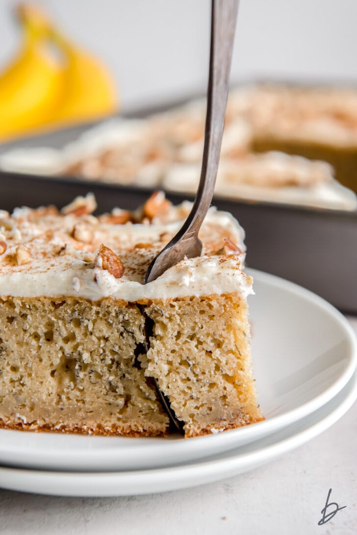 square slice of banana cake with cream sheet frosting and a fork sticking out of the corner