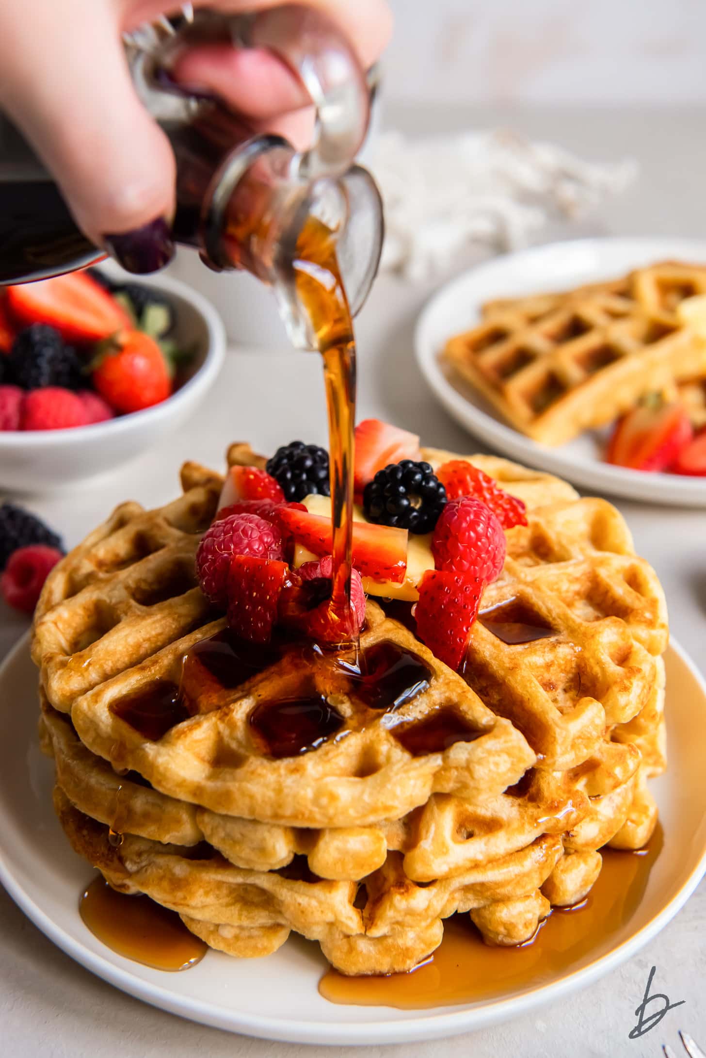 hand holding glass pitcher of maple syrup and pouring syrup over stack of waffles with berries