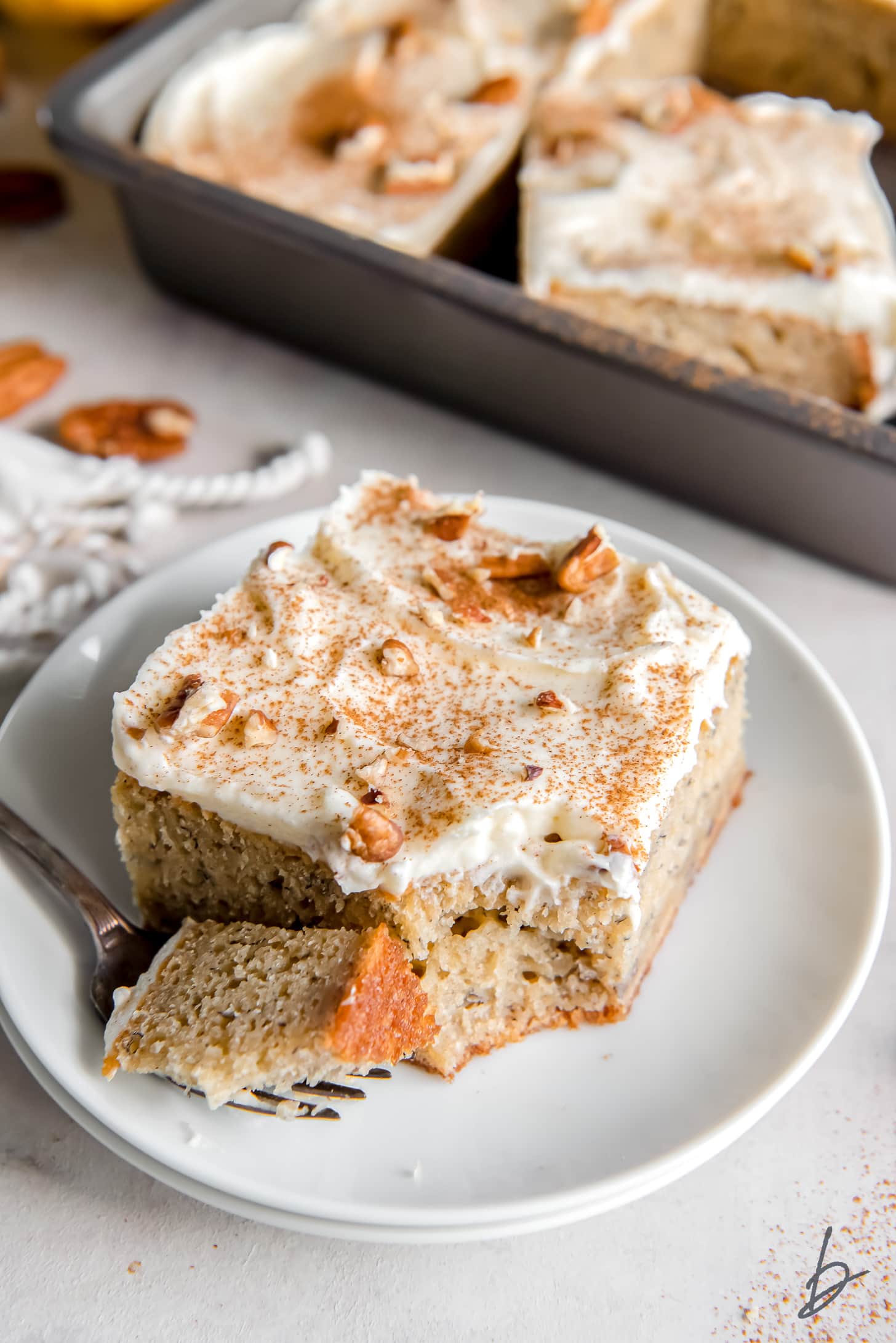 slice of banana sheet cake with cream cheese frosting and cinnamon with a fork holding a bite on a plate