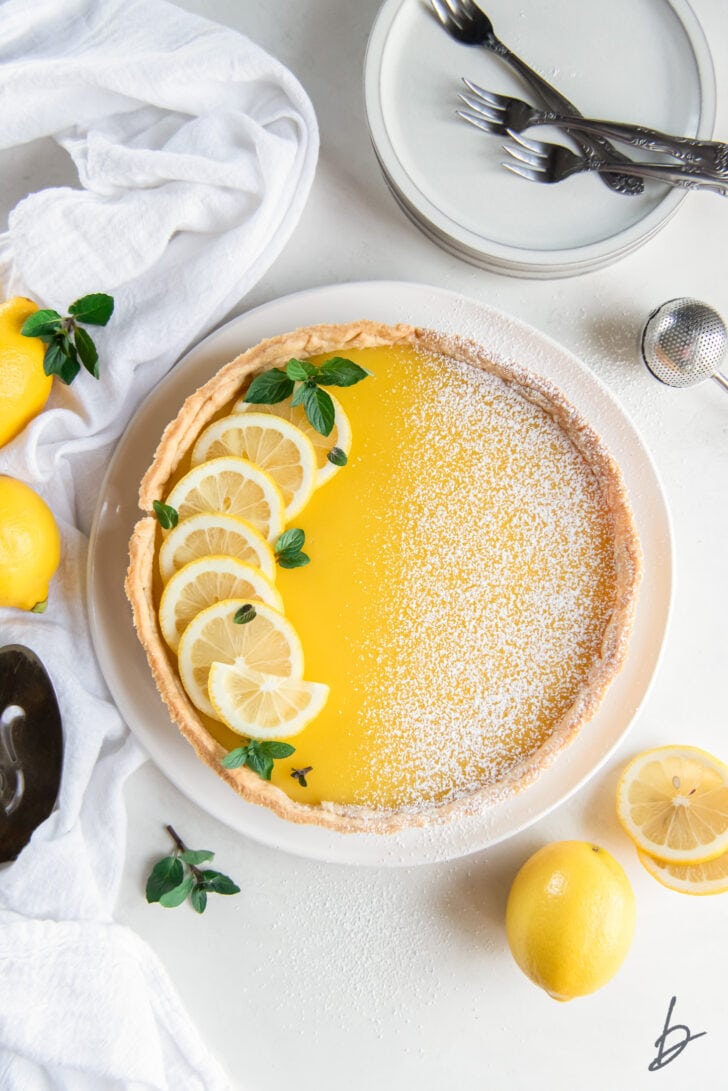 lemon curd tart garnished with lemon slices and dusting of confectioners sugar on a plate next to lemon halves, dessert forks and a kitchen cloth