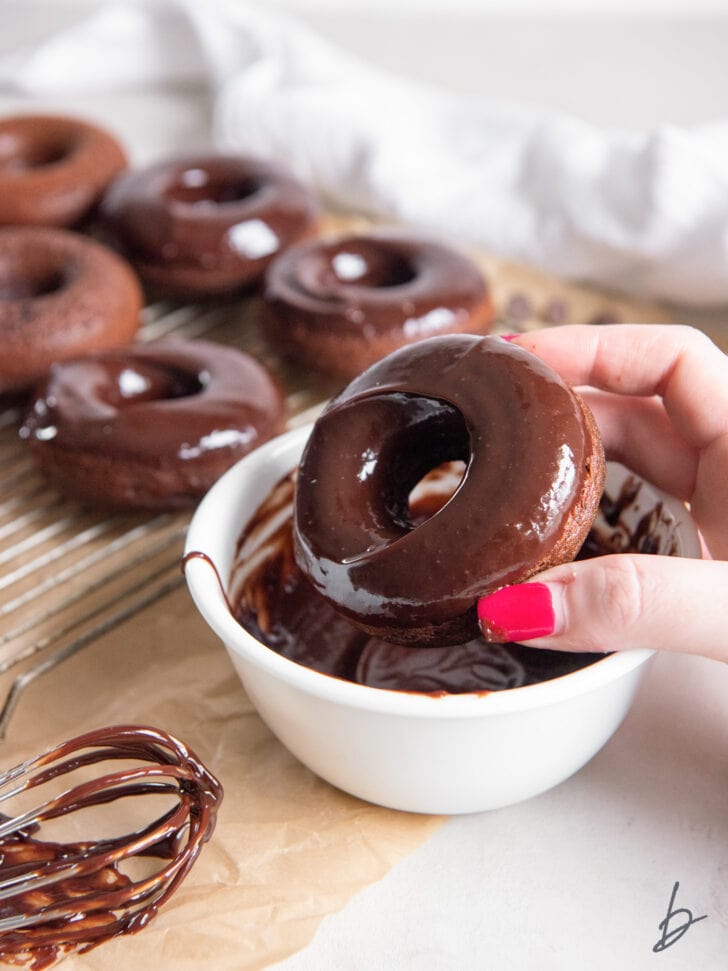hand holding chocolate donut with chocolate glaze over white bowl of melted chocolate
