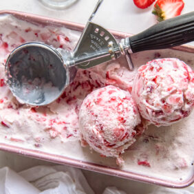 ice cream scoop sitting in 9x5 loaf pan of strawberry ice cream with two scoops sitting on top of ice cream