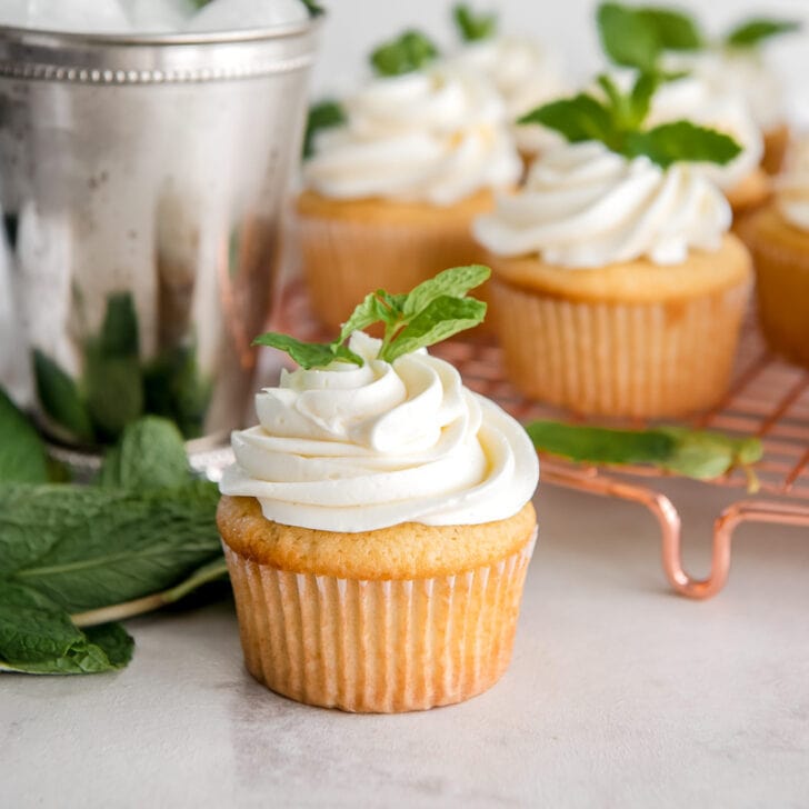 mint julep cupcake with frosting and mint leaf in front of more cupcakes and mint julep cup