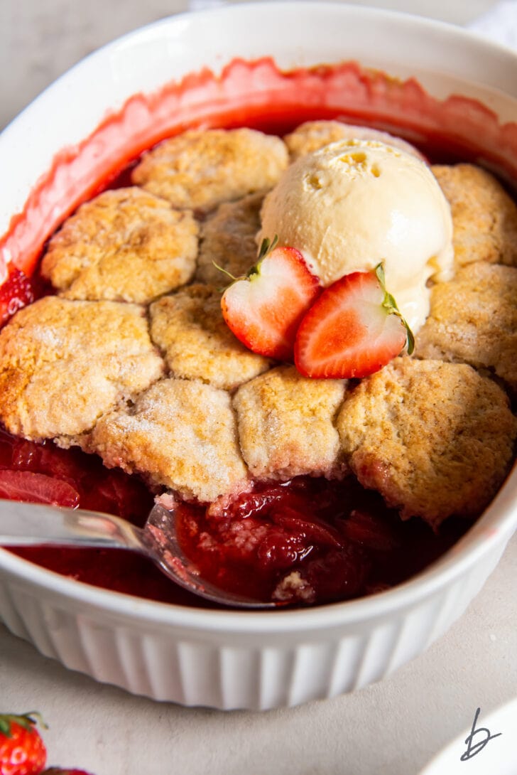 serving spoon in baking dish with strawberry cobbler and scoop of vanilla ice cream
