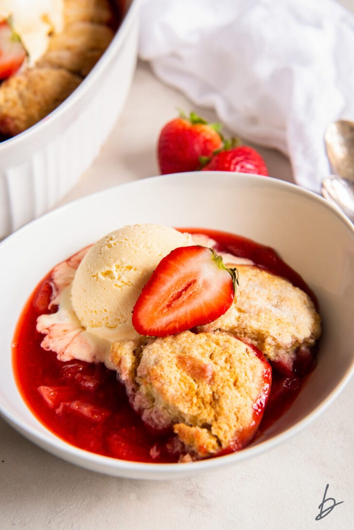 serving of strawberry cobbler in a bowl topped with a scoop of vanilla ice cream and fresh strawberry half