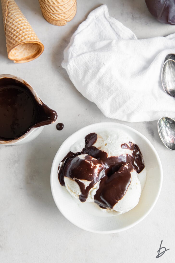 bowl of vanilla ice cream with hot fudge sauce next to creamer jar with fudge sauce and white kitchen linen