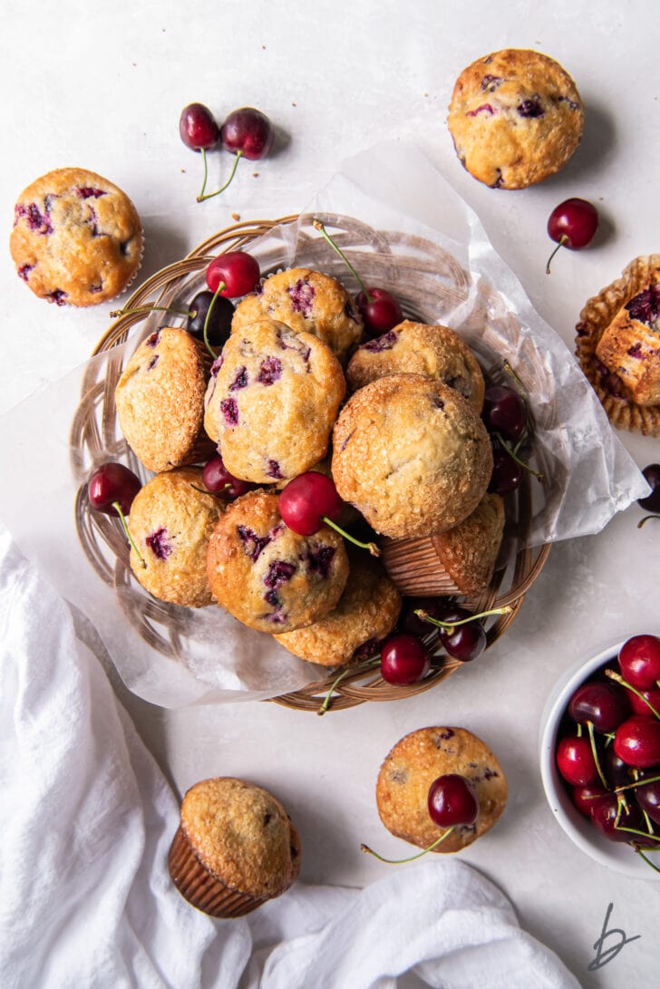 pile of cherry muffins on parchment paper in a basket next to a few more muffins