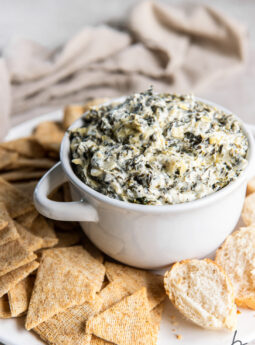 spinach artichoke dip in small white bowl with handles on tray with crackers