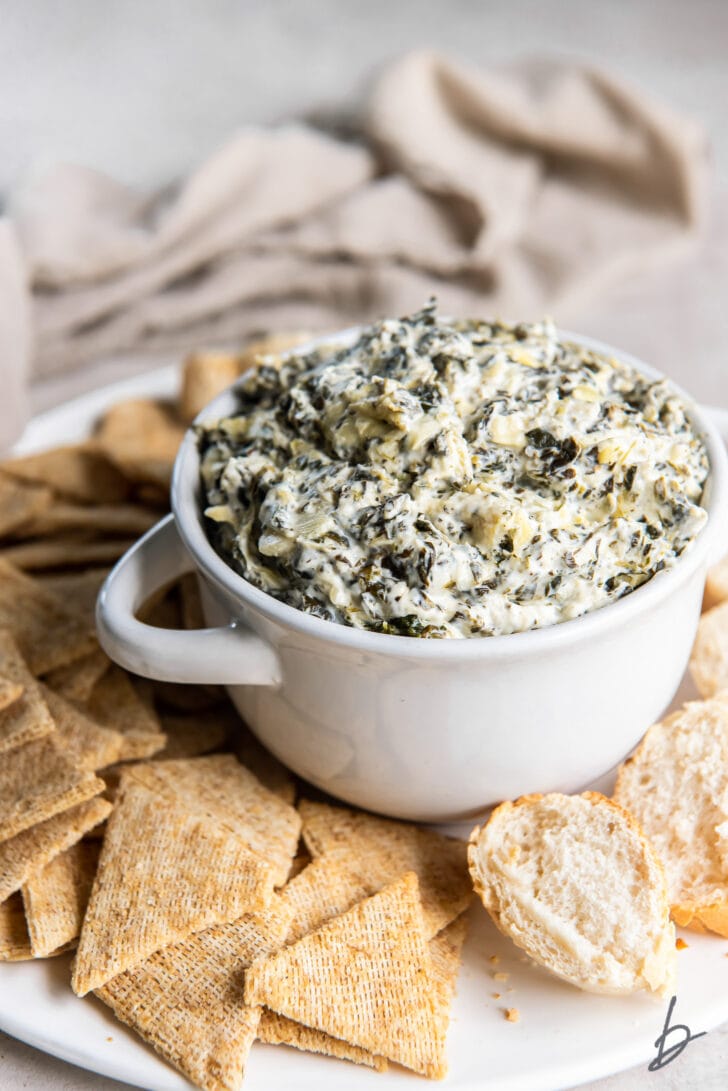 spinach artichoke dip in small white bowl with handles on tray with crackers
