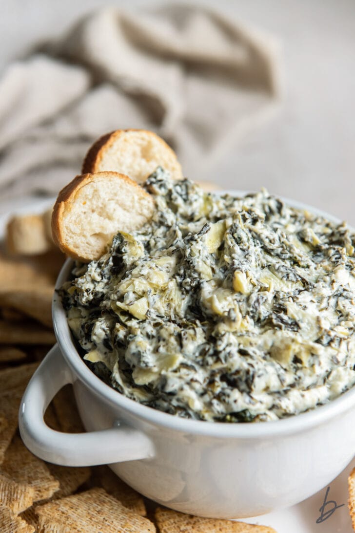 two pita chips in spinach artichoke dip in white bowl with handles