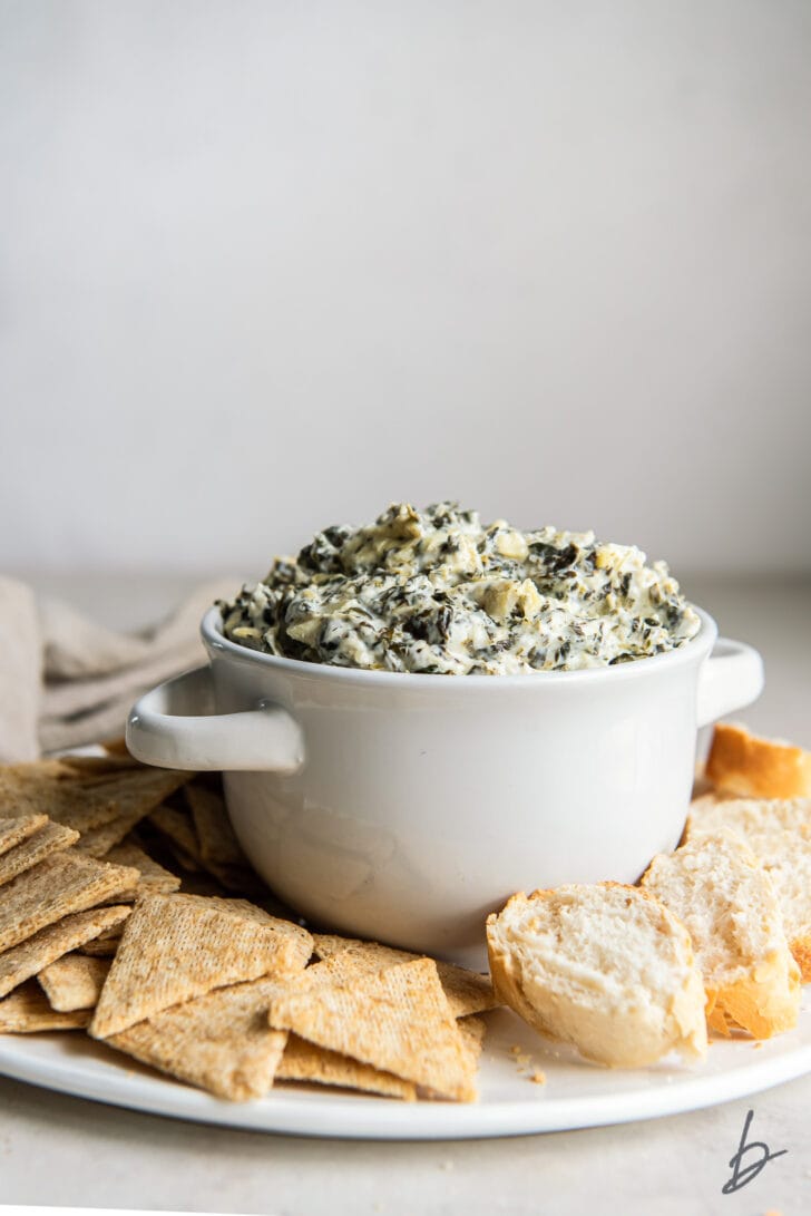 bowl of spinach artichoke dip on round platter with crostinis and crackers