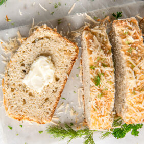 slice of parmesan herb quick bread with a pat of butter next to loaf with more slices and fresh herbs