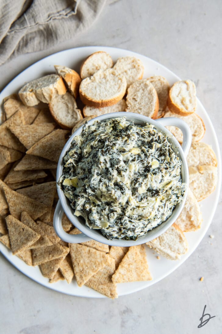 white round platter with crostinis and crackers surrounding bowl of spinach artichoke dip
