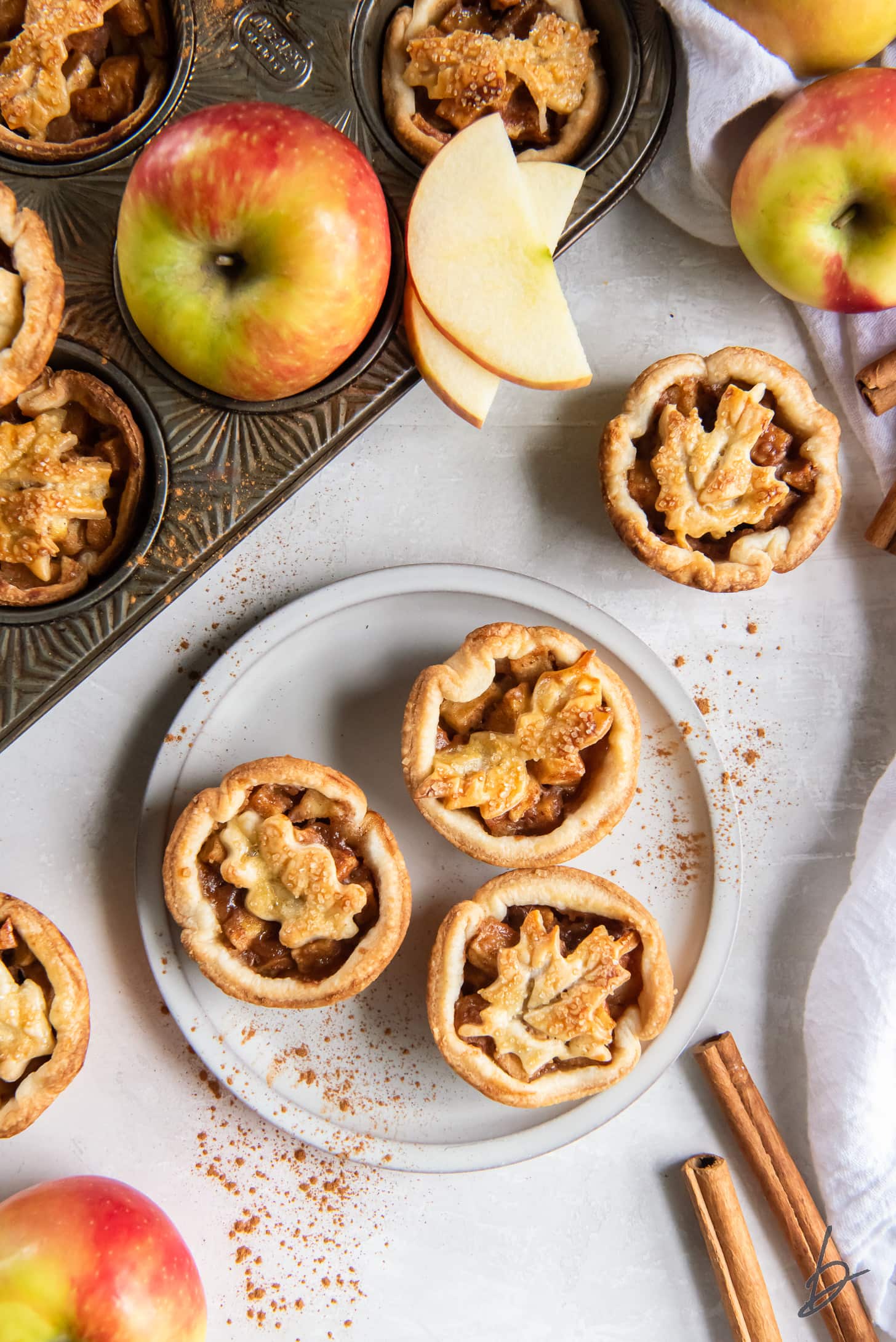 mini apple pies on plate next to apple slices and cinnamon sticks.