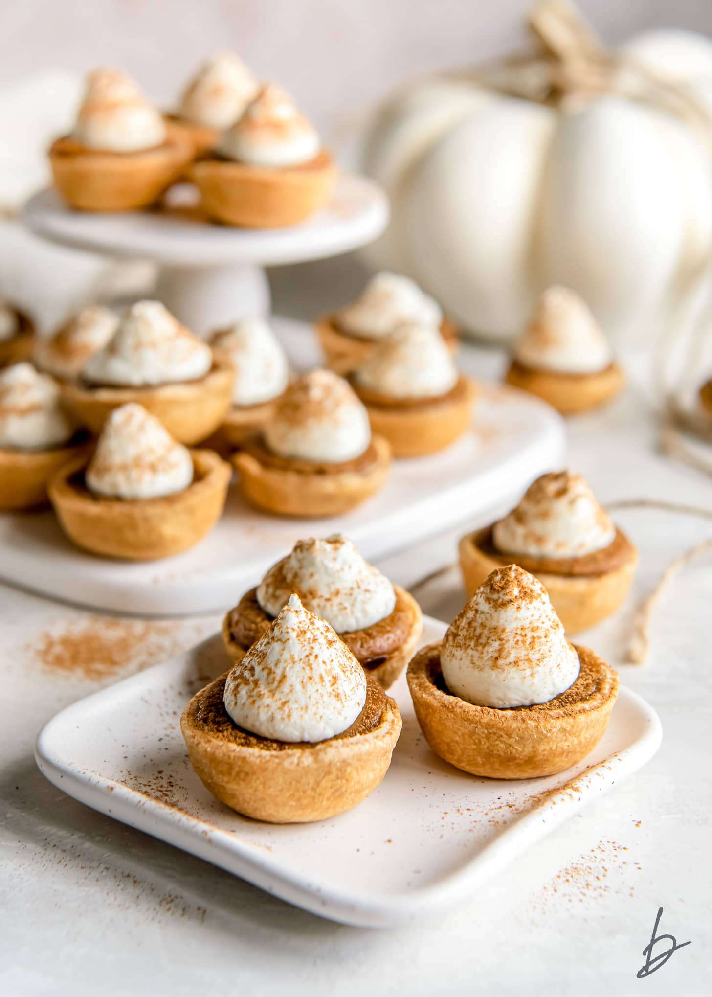 bite-sized pumpkin pies with dollops of whipped cream on plate in front of more pies on platter.