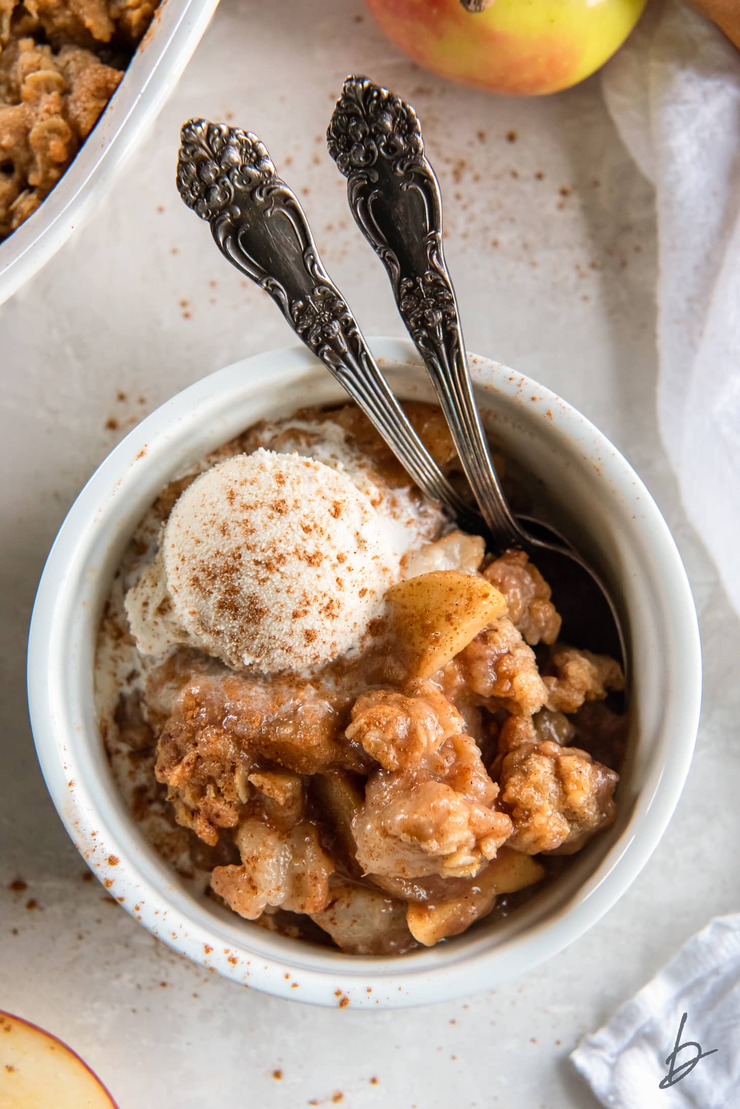 serving of apple pear crisp in a large ramekin with scoop of ice cream and two spoons.