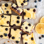 lemon blueberry cheesecake bars cut into squares next to lemon slices and fresh blueberries.