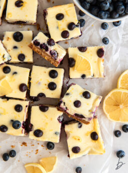 lemon blueberry cheesecake bars cut into squares next to lemon slices and fresh blueberries.