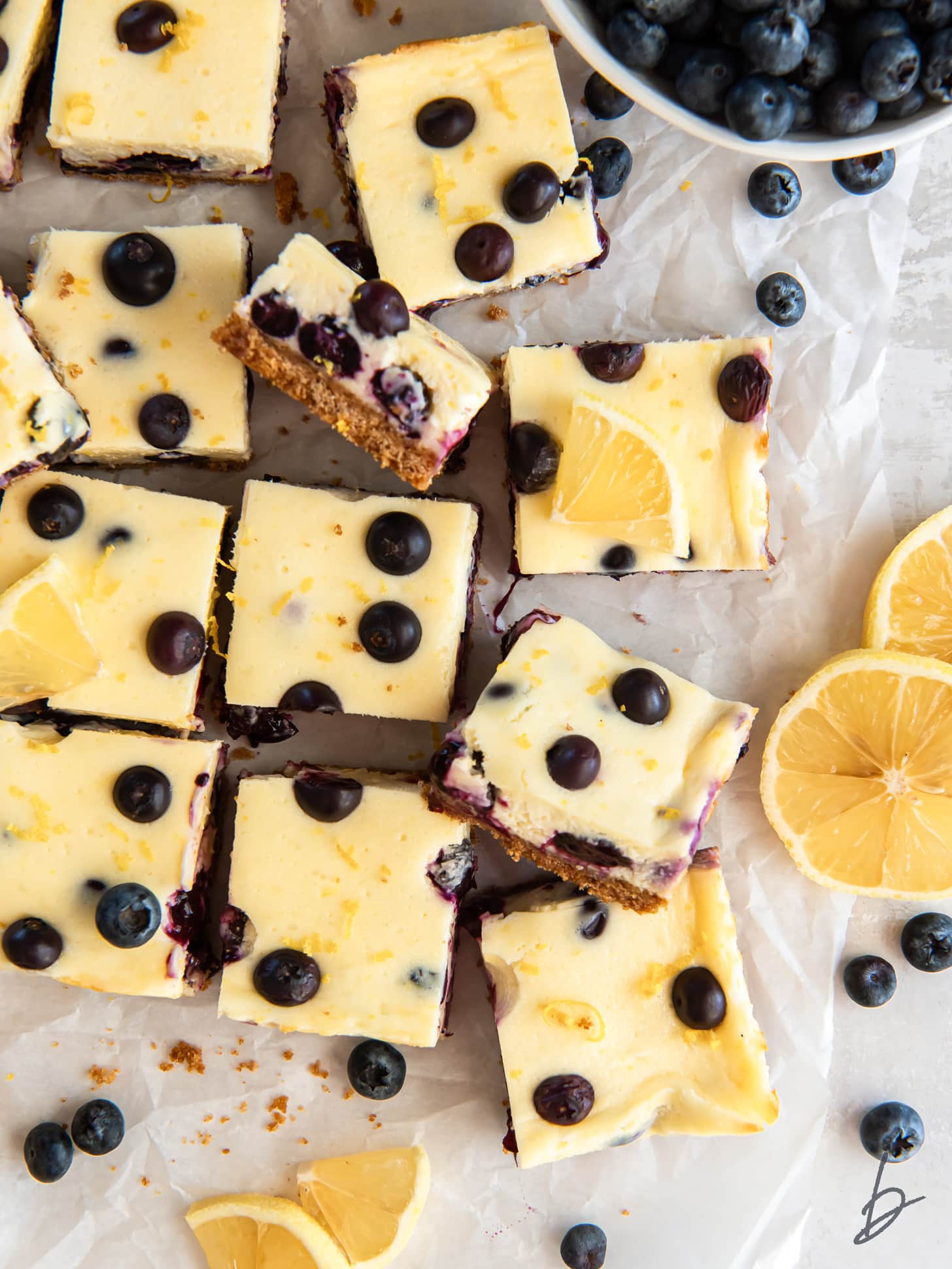 lemon blueberry cheesecake bars cut into squares next to lemon slices and fresh blueberries.