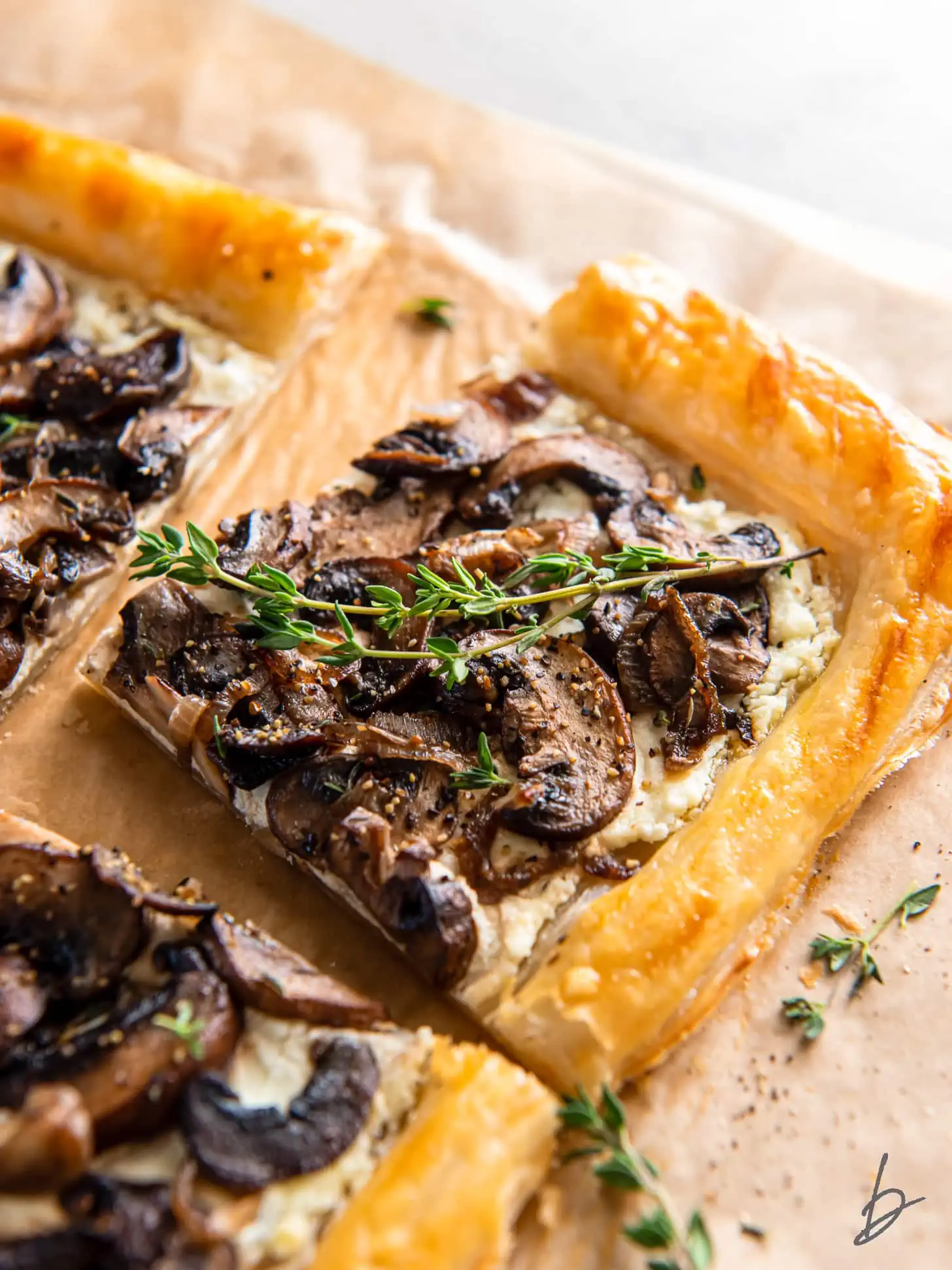 Rustic puff pastry tart with golden flaky edges showing visible layers, topped with caramelized mushrooms in browns and golds, dollops of creamy white goat cheese, scattered fresh thyme leaves, and a balsamic glaze drizzle creating dark amber streaks across the top.