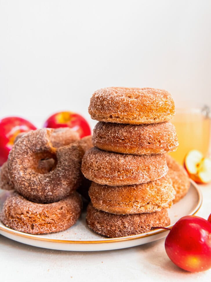stack of five baked apple cider donuts on a plate with more donuts.