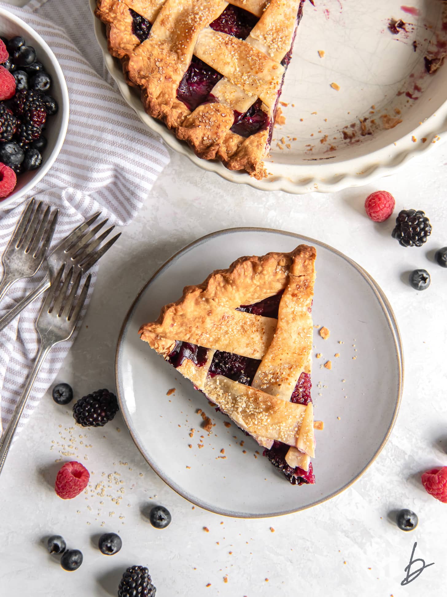 slice of triple berry pie on a round plate next to forks, pie plate and fresh berries.