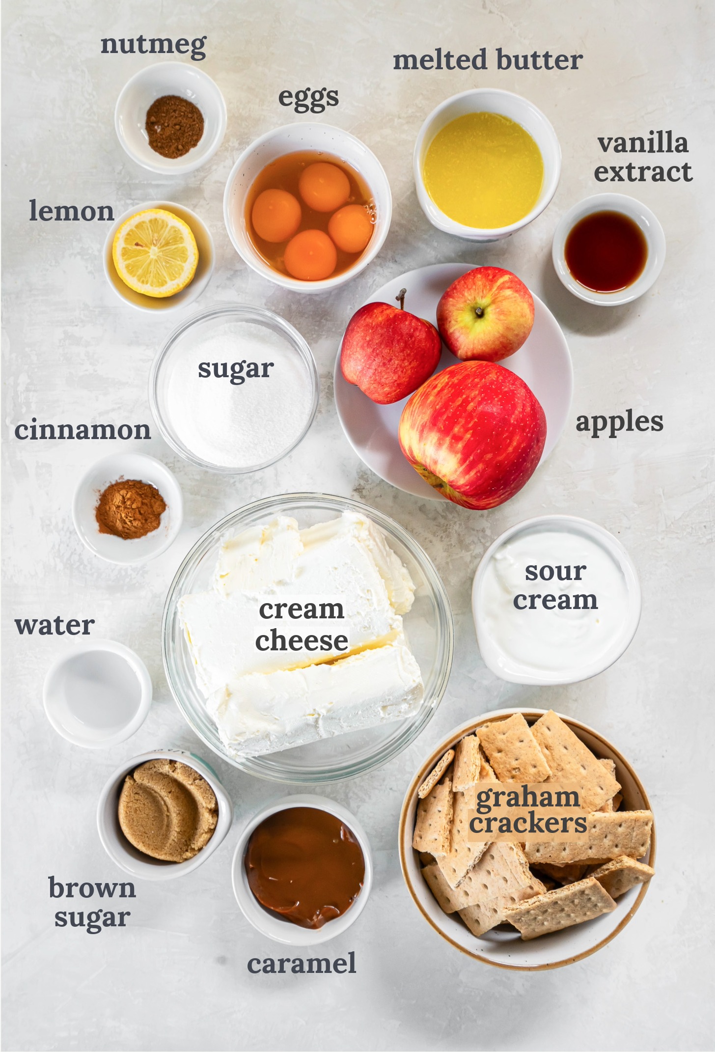 Apples near bowls of graham crackers, eggs, melted butter, sour cream, cream cheese and spices.