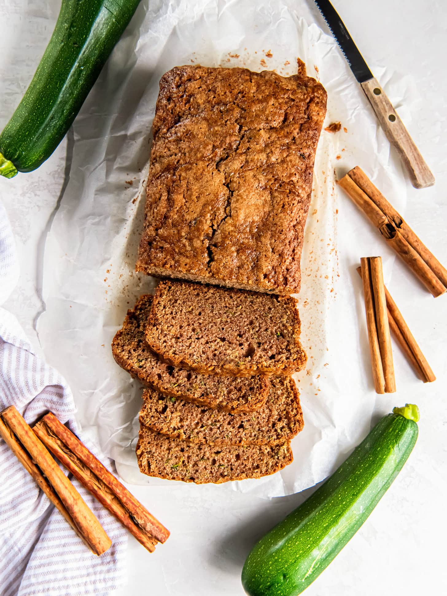tabletop with zucchini squash, cinnamon sticks and loaf of cinnamon zucchini bread with slices cut off the end.