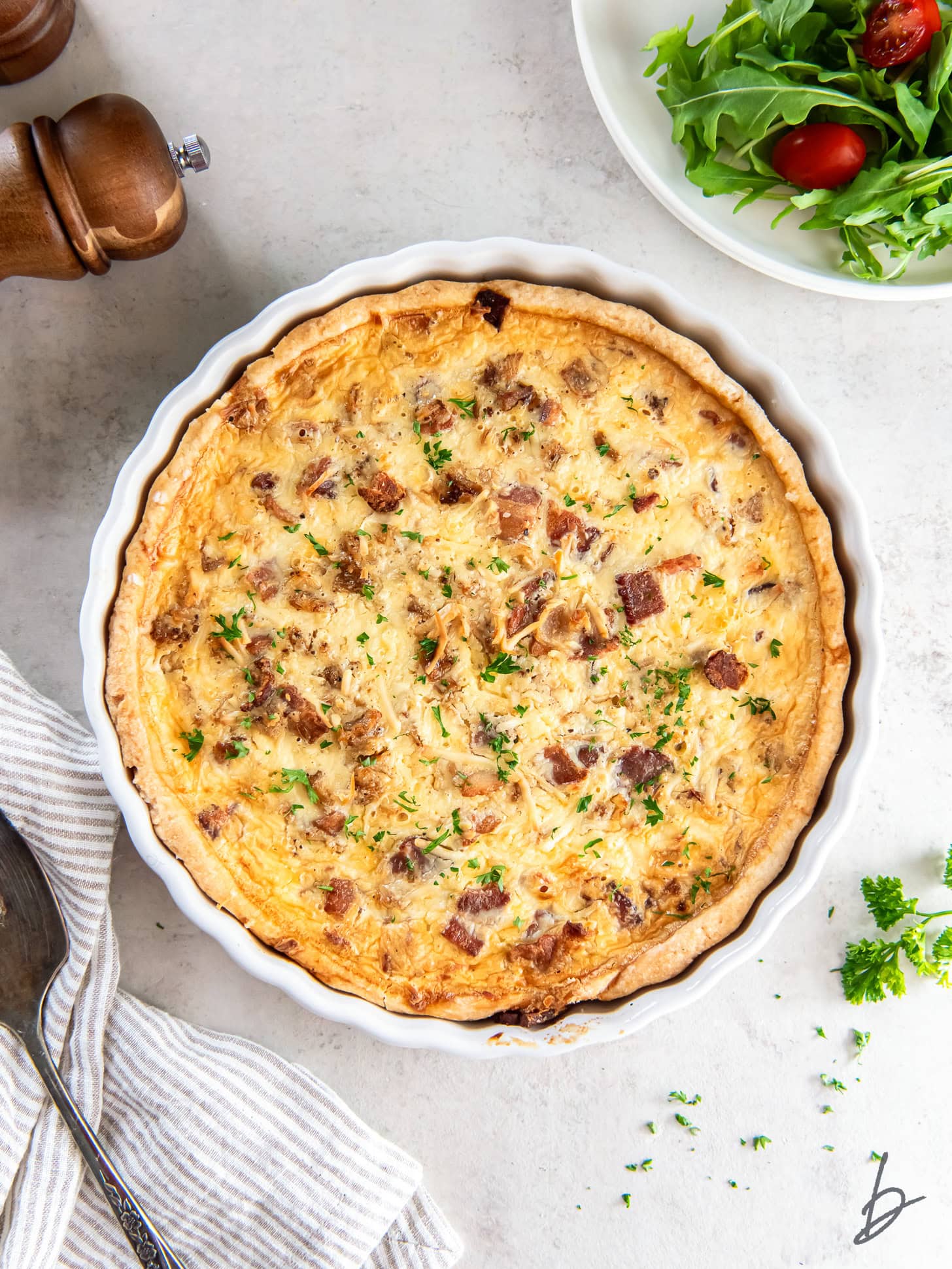 quiche lorraine on tabletop next to plate of fresh salad greens and striped kitchen towel.