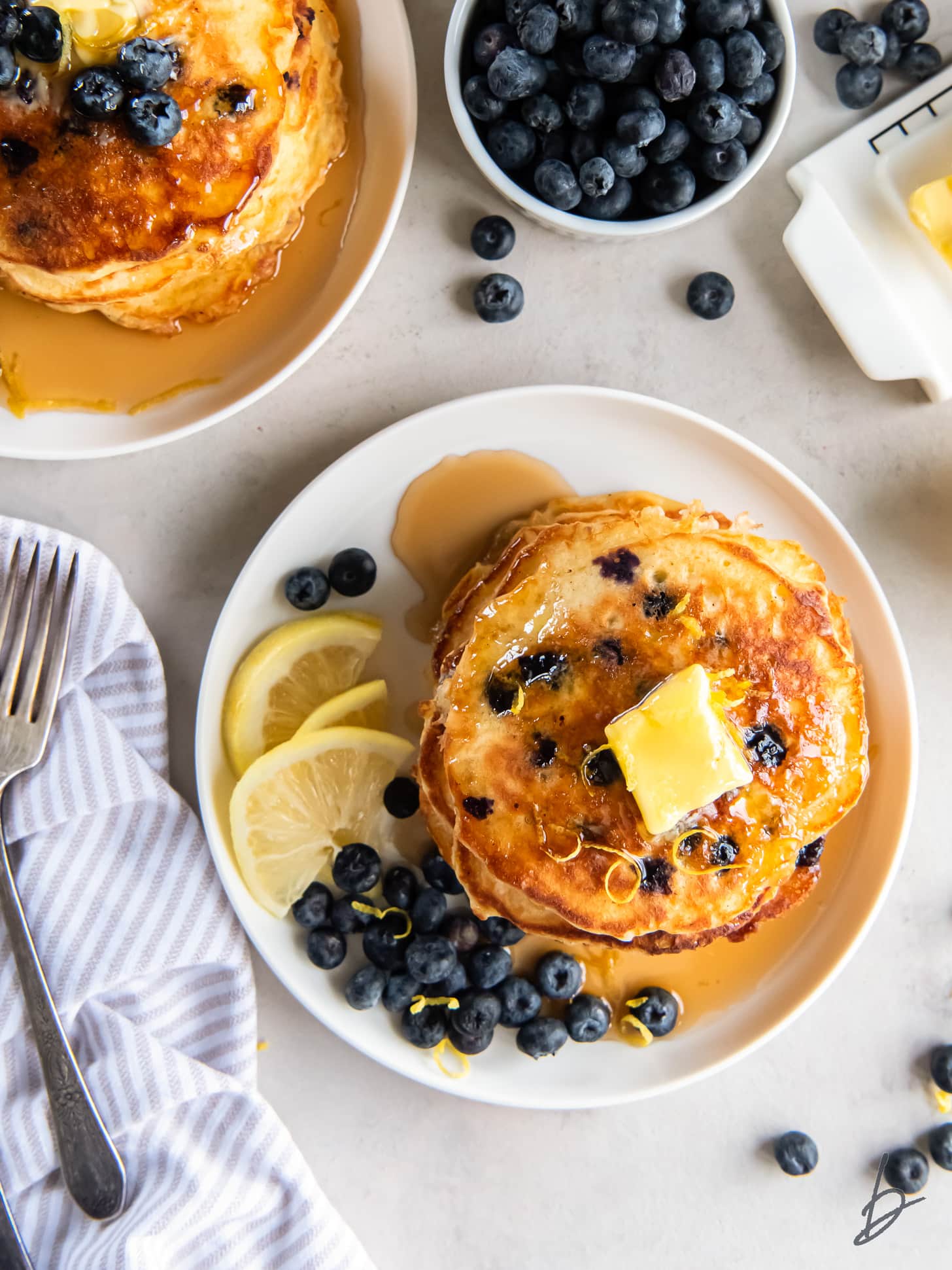 tabletop with plate of stack of lemon blueberry pancakes with fresh blueberries and lemon slices. 