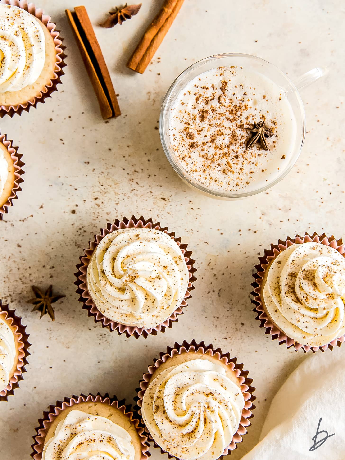 eggnog cupcakes on beige tabletop with cinnamon sticks and mug of eggnog next to cupcakes.